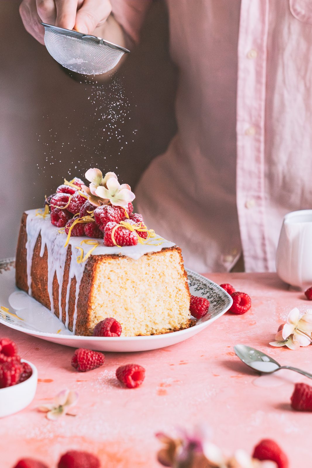 Mano espolvoreando azúcar glas sobre un cake de limón con frambuesas